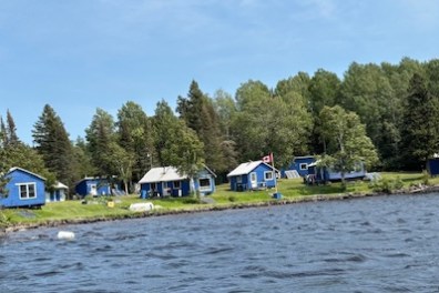Blue cottages by a lake with trees in the background.