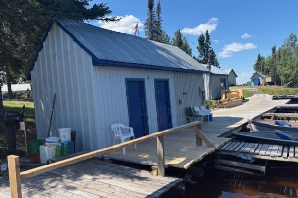 Wooden dock with small shed, chairs, and trees under a clear blue sky.