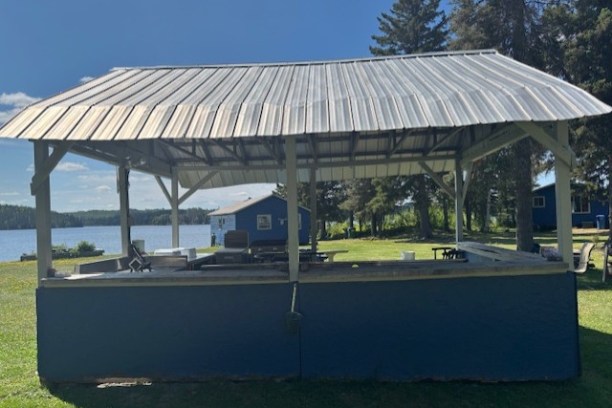 Open pavilion near a lake with picnic tables and blue cabins in the background.